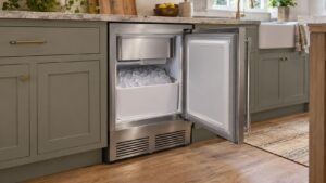A stainless steel 15-inch undercounter ice machine built into a cabinet run in an American kitchen, its door open to show a bin of ice