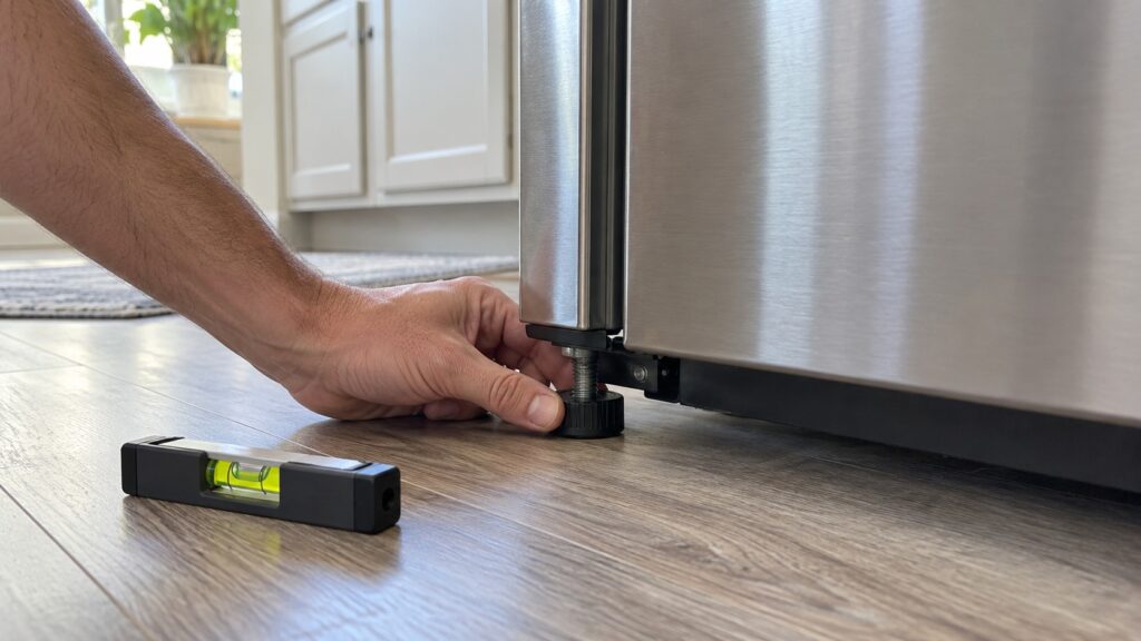 A hand adjusting a front leveling foot at the base of a refrigerator while a small level sits on the floor